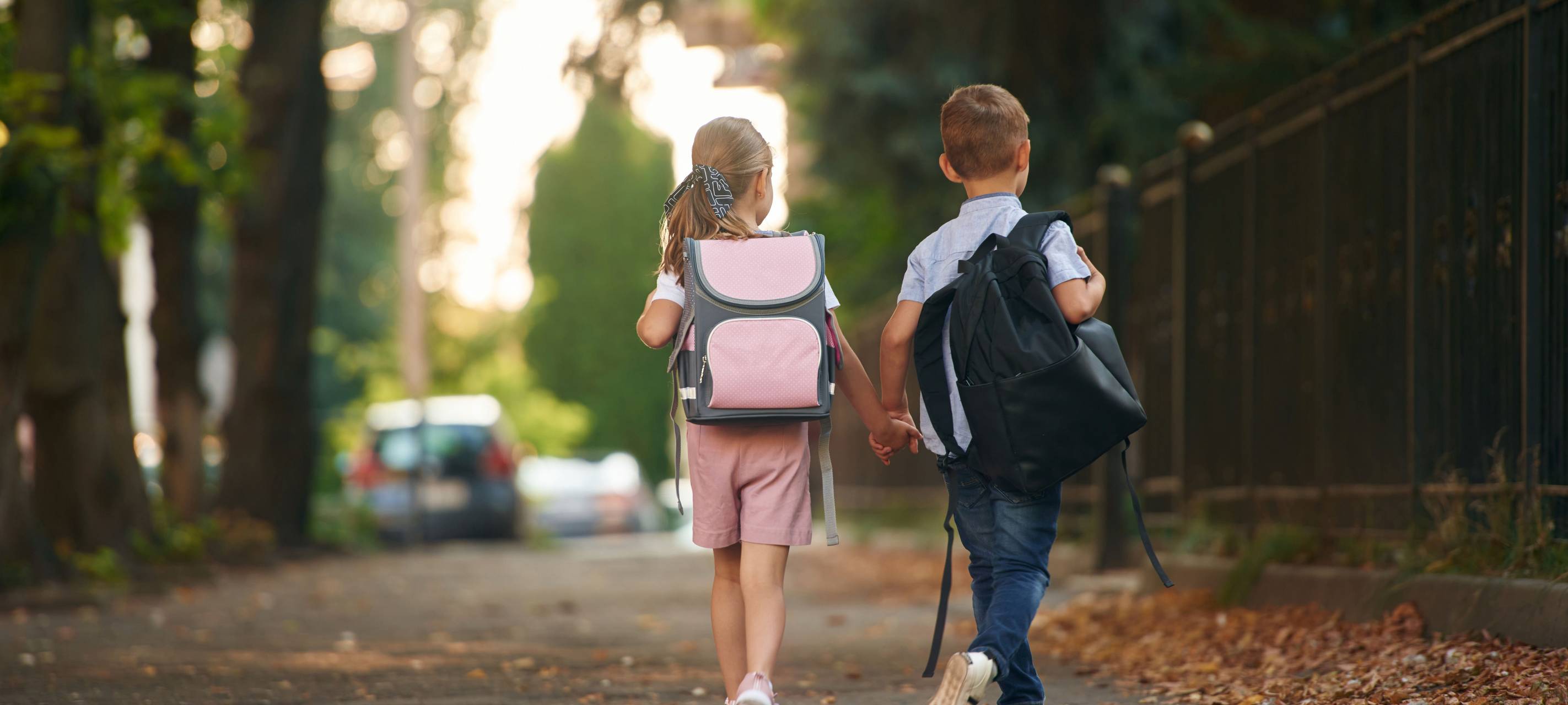 Ein Junge und ein Mädchen, mit Schulranzen auf dem Rücken halten sich an den Händen und laufen auf dem Gehweg
